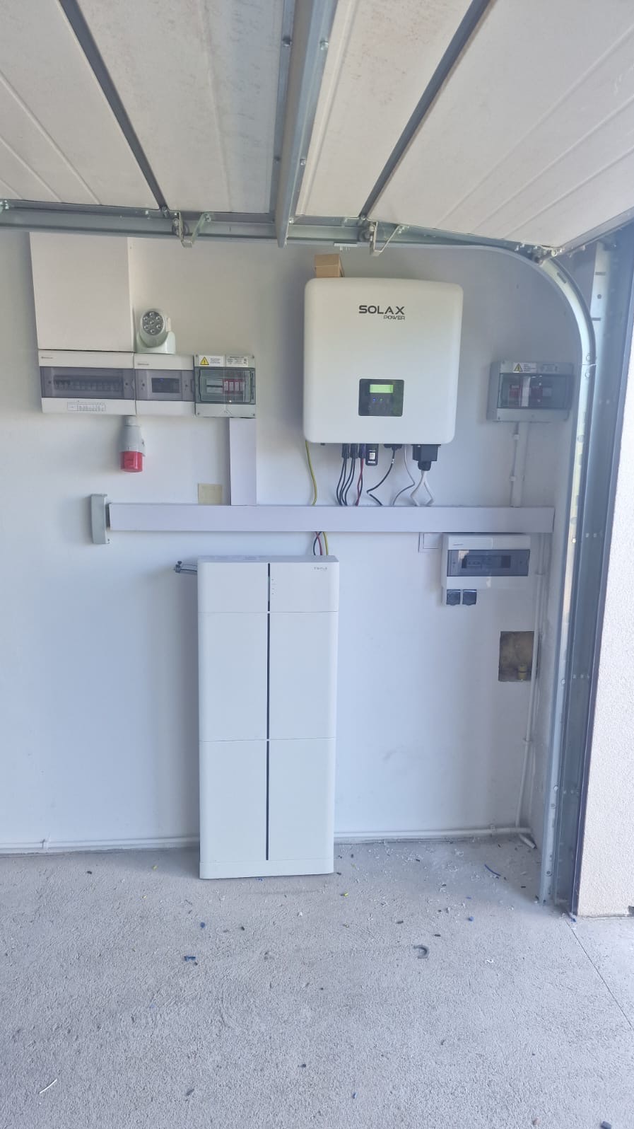 Biały magazyn energii z falownikiem Solax w garażu, widoczne zabezpieczenia elektryczne i okablowanie. Minimalistyczny design, surowe wykończenie podłogi.