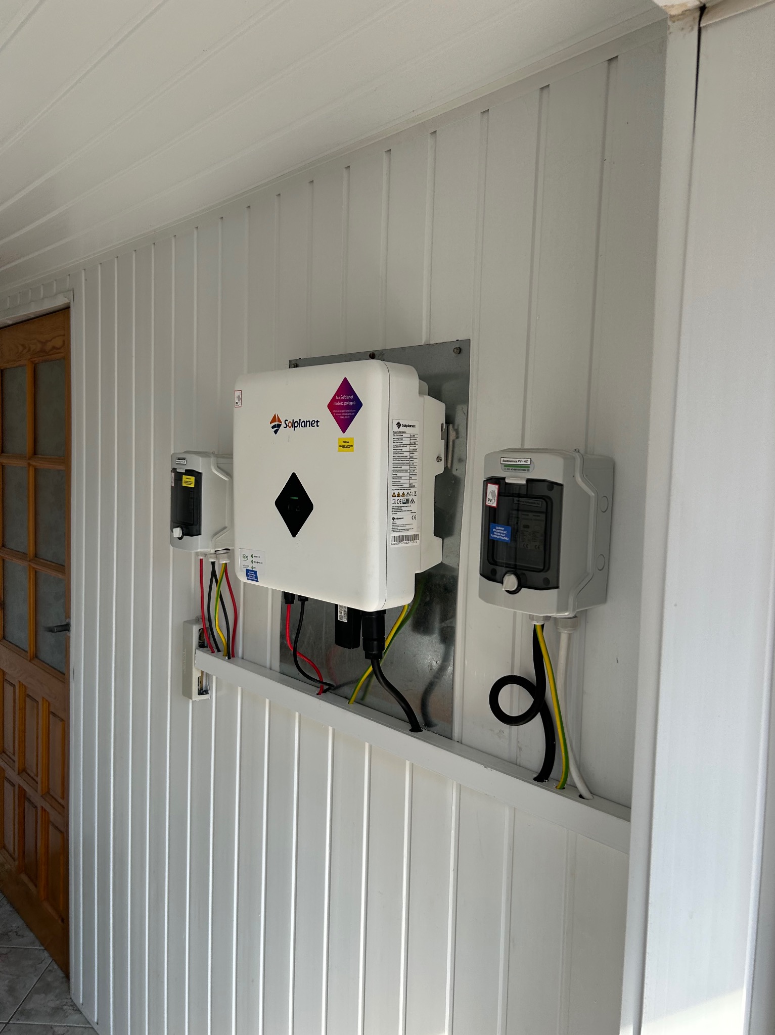 Biały inwerter Solplanet z okablowaniem i zabezpieczeniami zamontowany na białej, panelowej ścianie wewnątrz budynku, obok drewnianych drzwi.