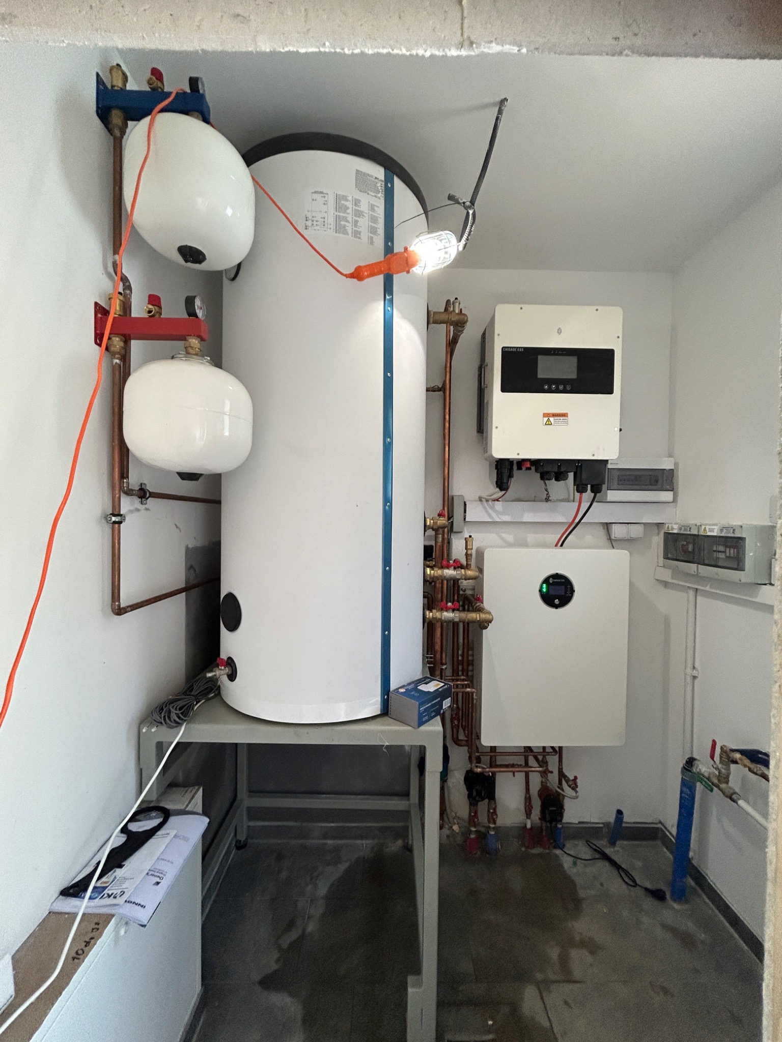 Biały zbiornik buforowy na metalowym stelażu, obok inwerter fotowoltaiczny i instalacja z miedzianych rur w pomieszczeniu z surowym betonowym wykończeniem.