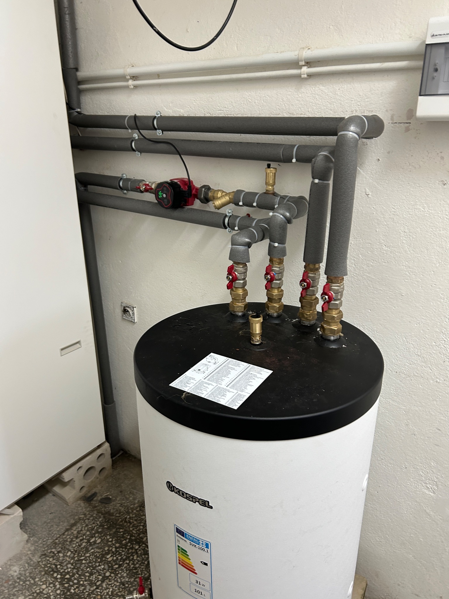 Bojler Kospel z podłączonymi rurami i zaworami, widoczne izolacje termiczne na rurach. Na górze bojlera leży instrukcja obsługi. Szare rury na ścianie.