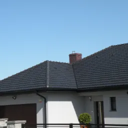 Nowoczesny dom jednorodzinny z ciemnoszarą dachówką modułową i ceglanym kominem z metalowym zakończeniem, widok z dołu na tle błękitnego nieba.