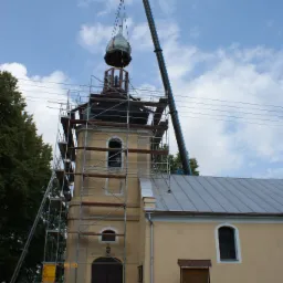 Montaż kopuły na wieży kościoła z użyciem dźwigu, rusztowanie wokół wieży, błękitne niebo w tle.
