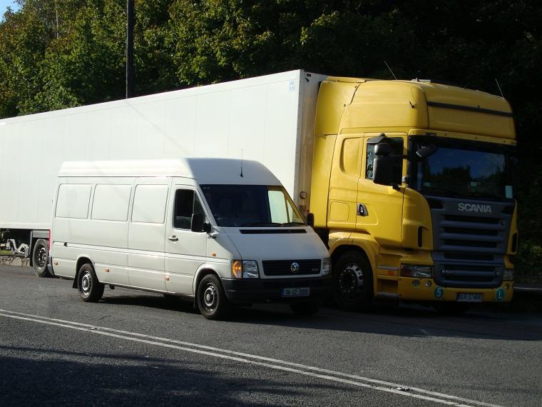 Biały bus dostawczy obok żółtej ciężarówki Scania na tle zieleni. Transport drogowy, widok z przodu, światło dzienne. Tablice rejestracyjne widoczne.