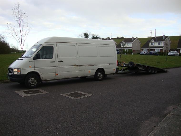 Biały bus z przyczepą lawetą na tle domów jednorodzinnych i pochmurnego nieba. Pojazd zaparkowany na asfaltowej drodze.