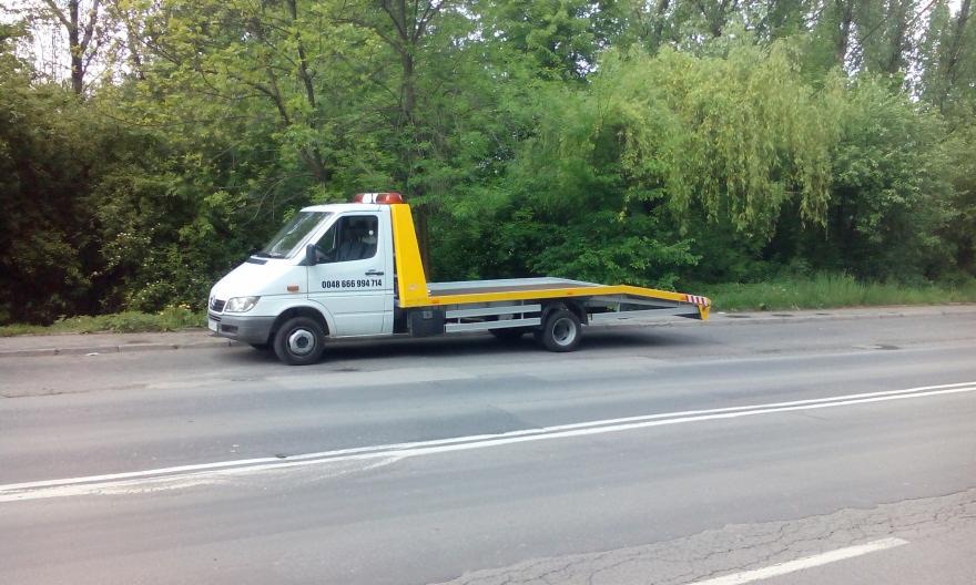 Biała laweta Mercedes Sprinter z żółtymi elementami na poboczu drogi. Widoczny numer telefonu na kabinie, zielone drzewa w tle. Dzień, lekko pochmurno.