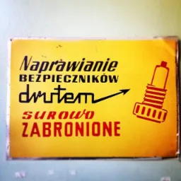 Żółta tabliczka z napisem 'Naprawianie bezpieczników drutem surowo zabronione' i schematycznym rysunkiem bezpiecznika.