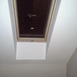 Okno dachowe z drewnianą ramą, zamontowane w białym suficie pod skosem, widok z dołu.