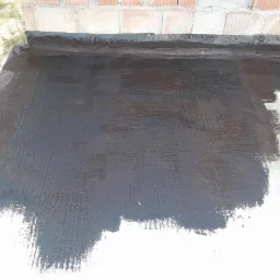 Świeżo zaizolowany, czarny dach płaski z widocznymi śladami pędzla i fragmentem muru z cegieł w tle.