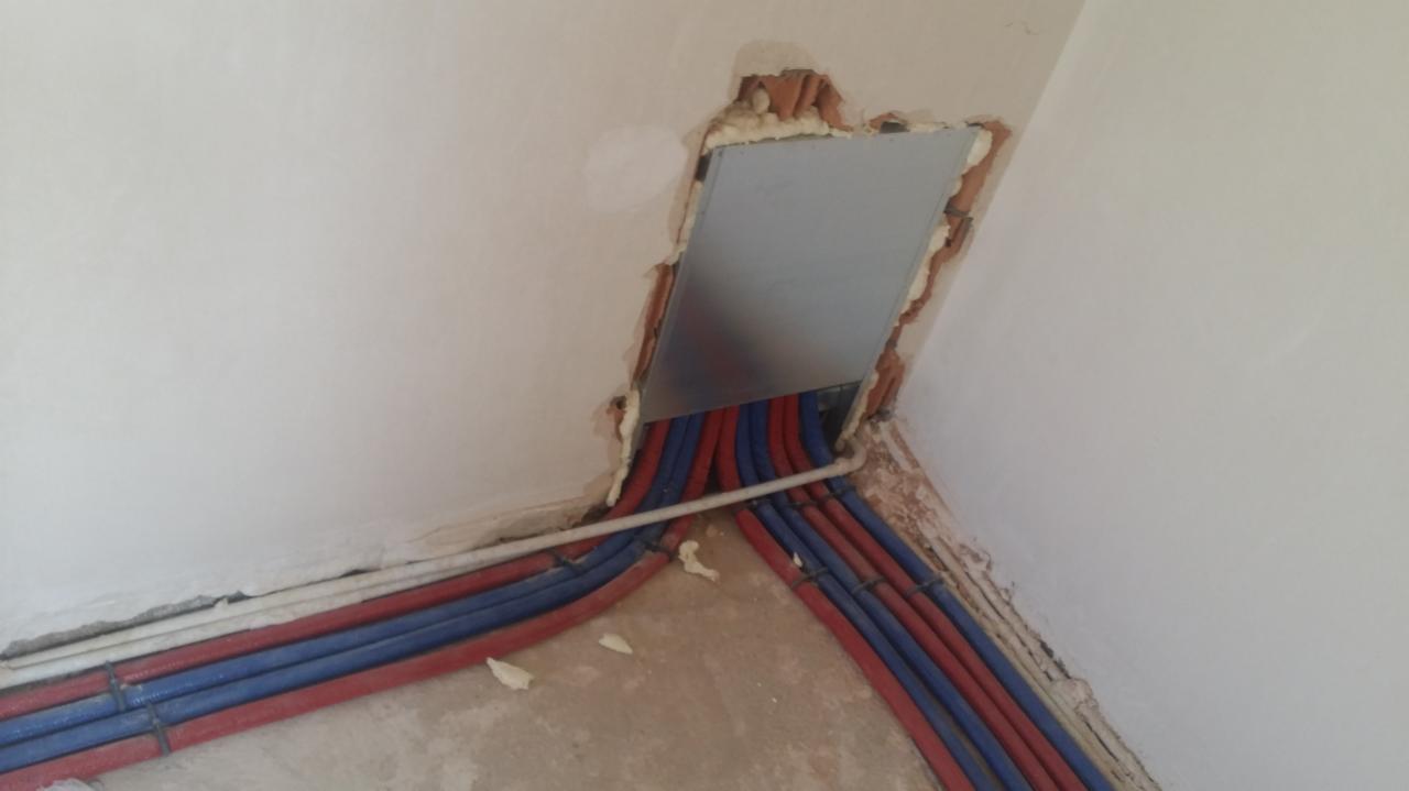 Instalacja hydrauliczna z czerwono-niebieskimi rurami wychodzącymi ze ściany, zabezpieczona pianką montażową wokół otworu rewizyjnego z szarą klapką.