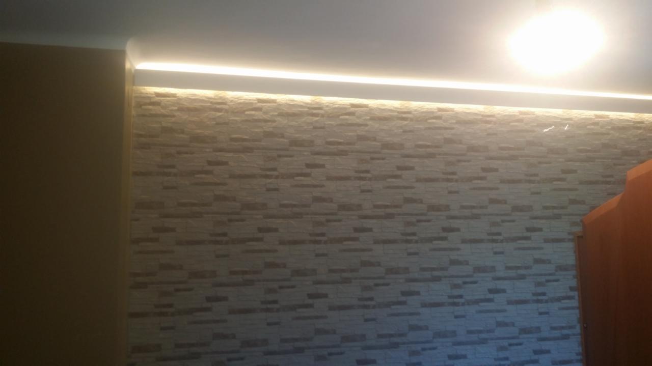 Ściana wykończona dekoracyjną okładziną imitującą kamień, oświetlona listwą LED pod sufitem, w ciepłej tonacji.