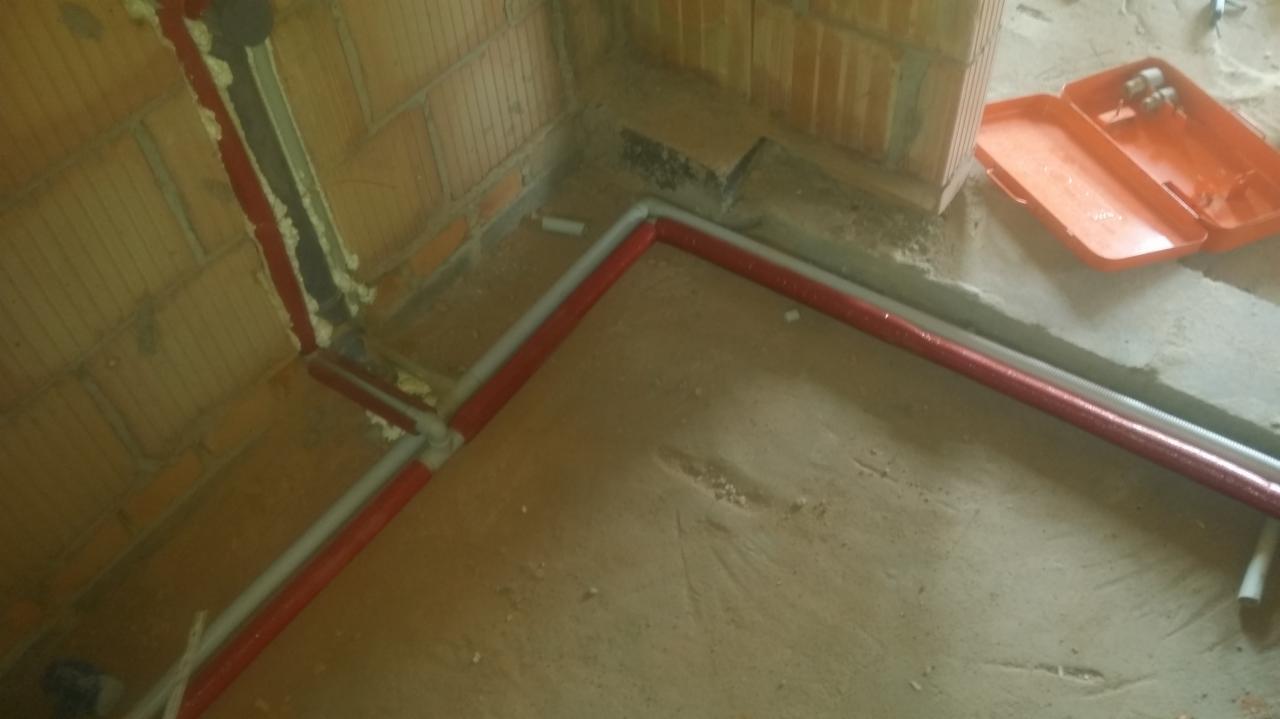 Wewnętrzny róg surowego pomieszczenia z instalacją wodną: czerwone i szare rury przytwierdzone do ceglanej ściany pianką montażową, pomarańczowa skrzynka z narzędziami w tle.