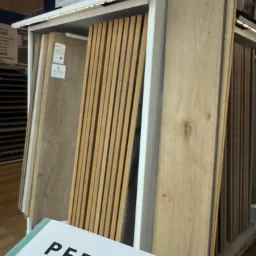 Ekspozycja paneli podłogowych Pergo w sklepie: różne odcienie drewna w pionowym stojaku z oświetleniem LED, widoczne próbki i logo producenta na pierwszym planie.