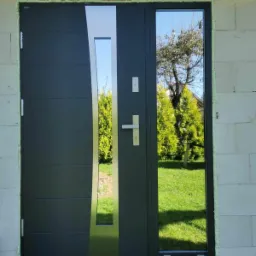 Grafitowe drzwi zewnętrzne z przeszkleniem i srebrnym panelem bocznym, zamontowane w surowym murze. Widoczna zieleń ogrodu w odbiciu szyby.