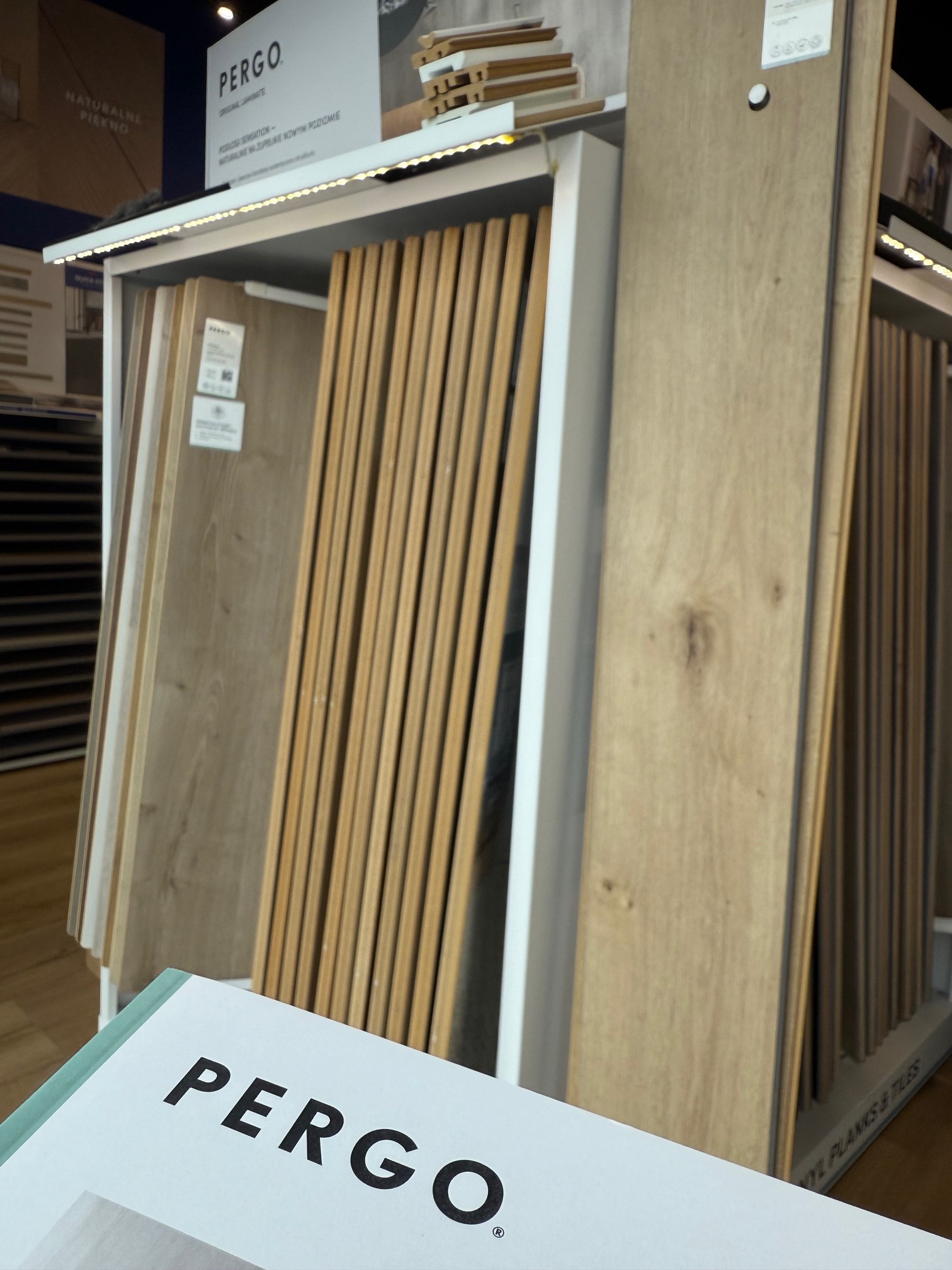 Ekspozycja paneli podłogowych Pergo w sklepie: różne odcienie drewna w pionowym stojaku z oświetleniem LED, widoczne próbki i logo producenta na pierwszym planie.