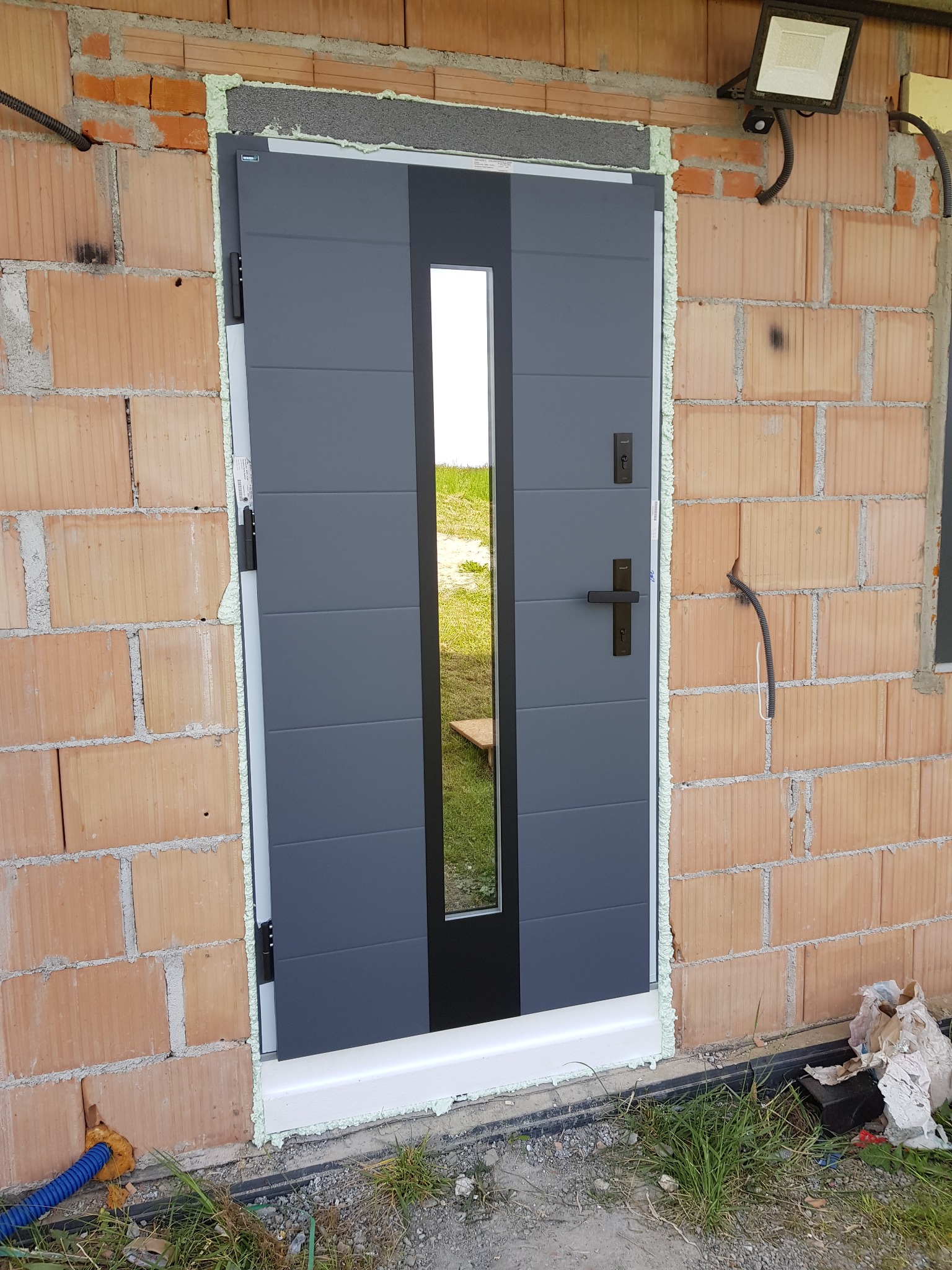 Montaż antracytowych drzwi zewnętrznych z przeszkleniem, widoczne pianowanie wokół ościeżnicy w surowym murze z pustaków ceramicznych. Widoczny reflektor.