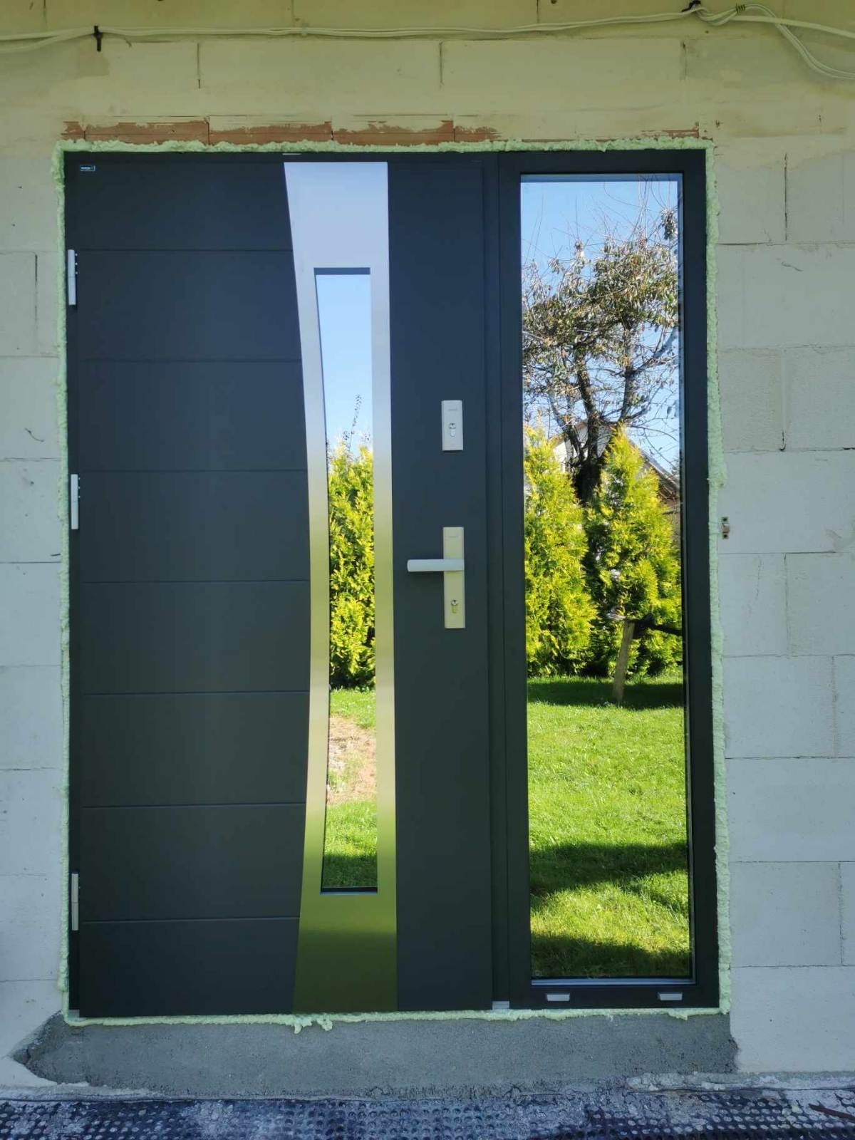 Grafitowe drzwi zewnętrzne z przeszkleniem i srebrnym panelem bocznym, zamontowane w surowym murze. Widoczna zieleń ogrodu w odbiciu szyby.