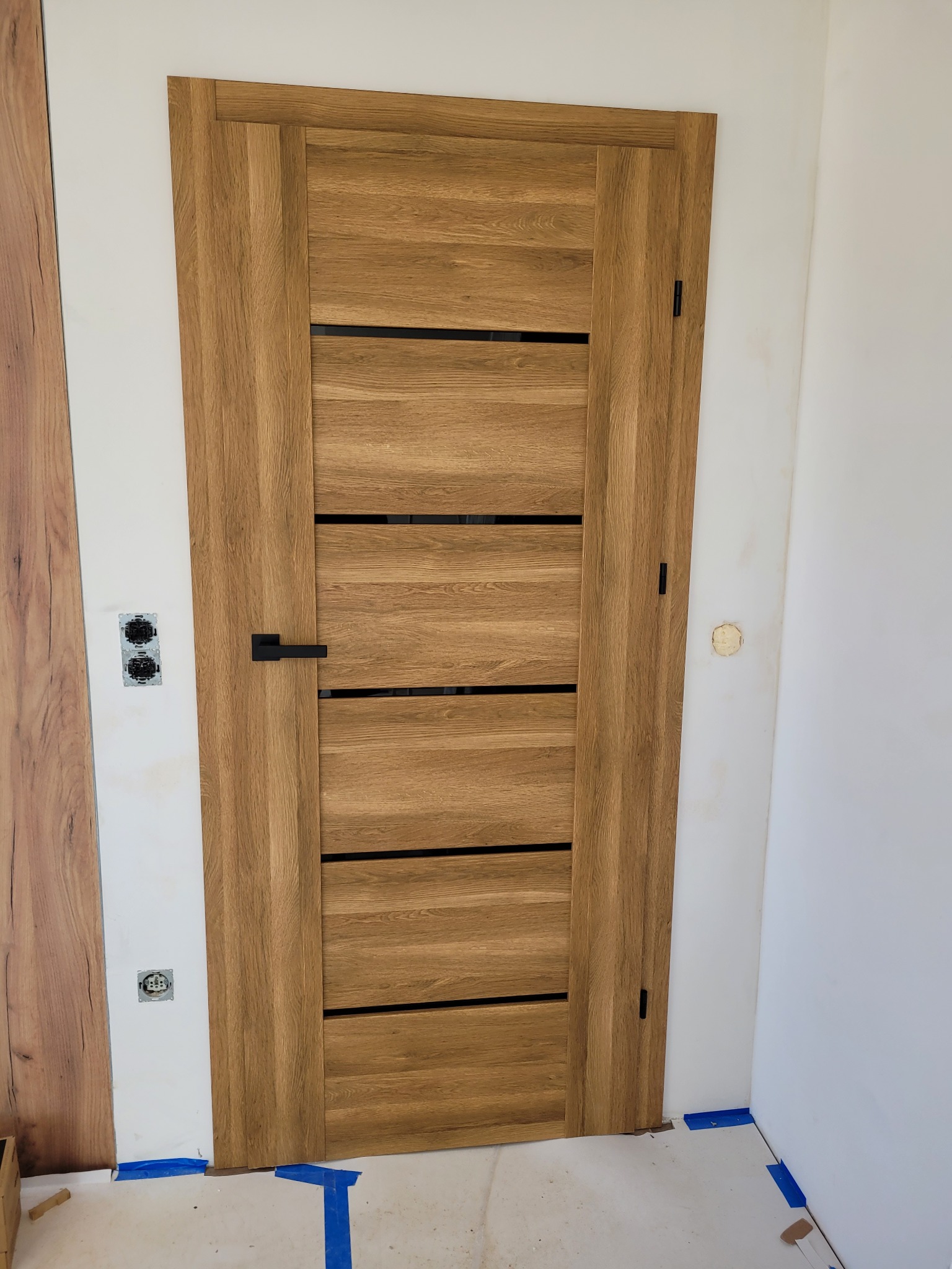 Dębowa okleina drzwi wewnętrznych z poziomymi, czarnymi intarsjami i czarną klamką, zamontowana w surowym wnętrzu. Widoczne ślady po pracach wykończeniowych.