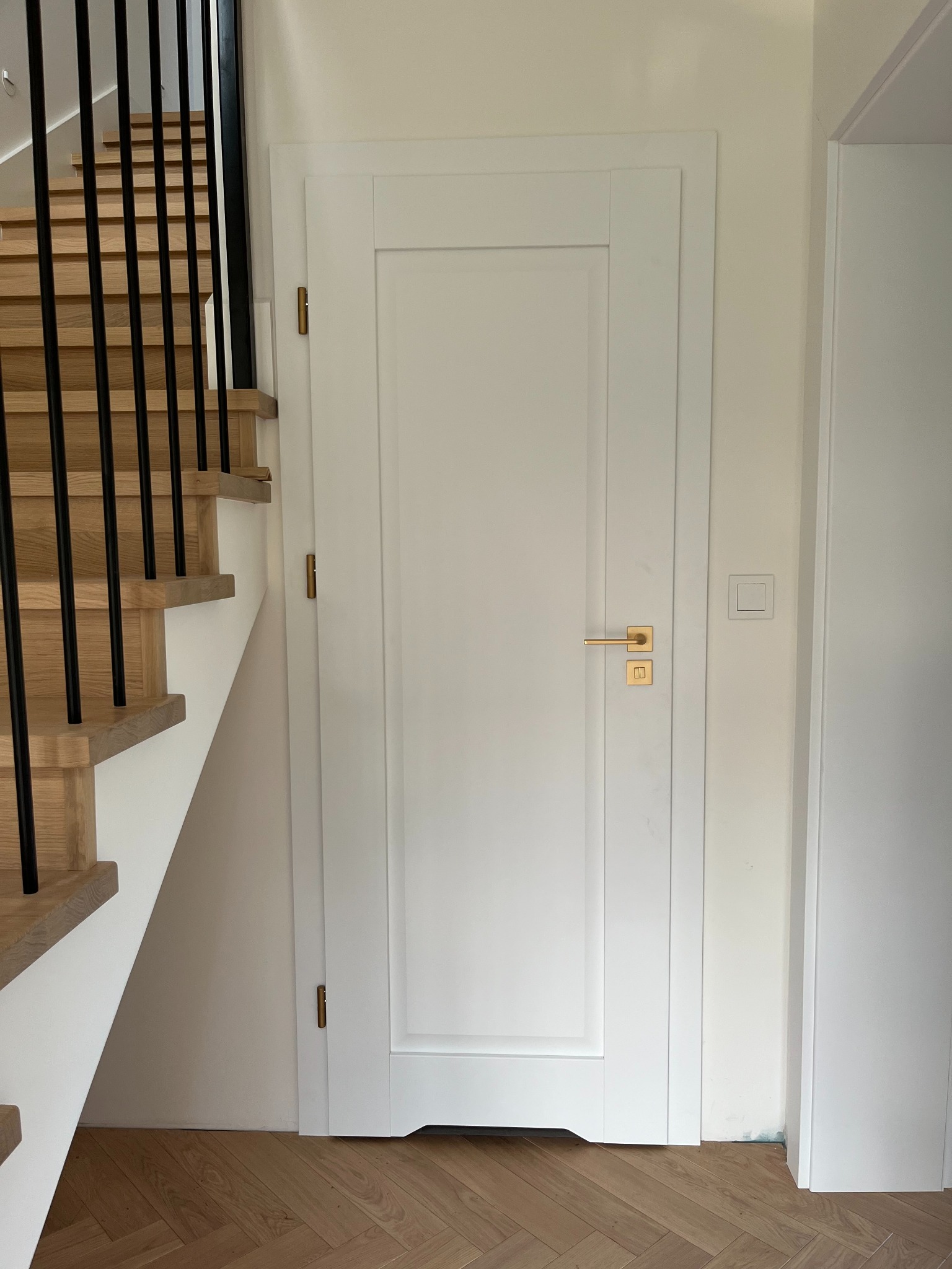 Białe drzwi wewnętrzne z prostokątnym panelem i złotą klamką, widoczne schody z drewnianymi stopniami i czarną balustradą, parkiet ułożony w jodełkę.
