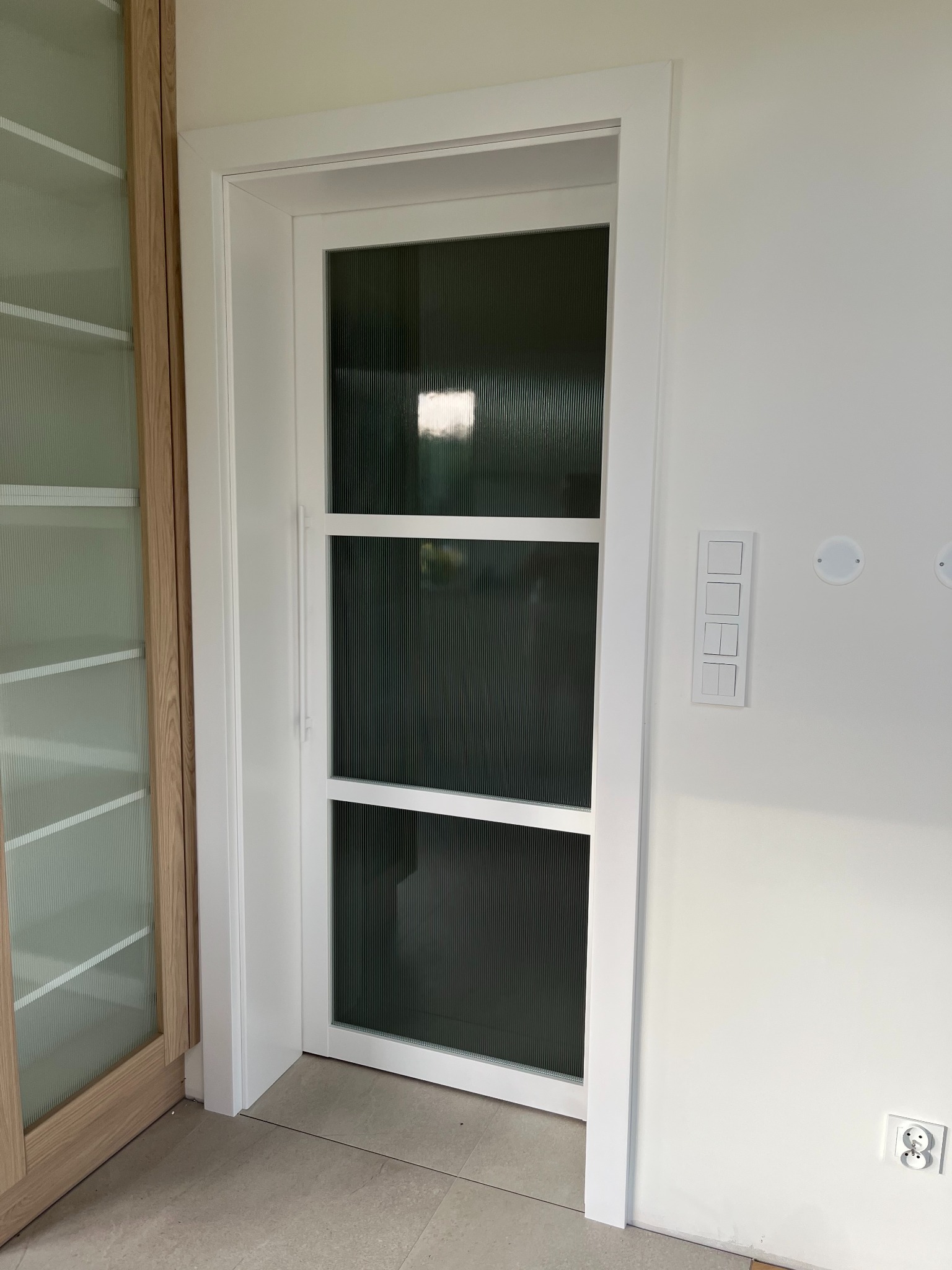 Białe drzwi wewnętrzne z trzema szybami o prążkowanej fakturze, osadzone w białej futrynie. Widoczne panele podłogowe i fragment szafy z przeszkleniami.