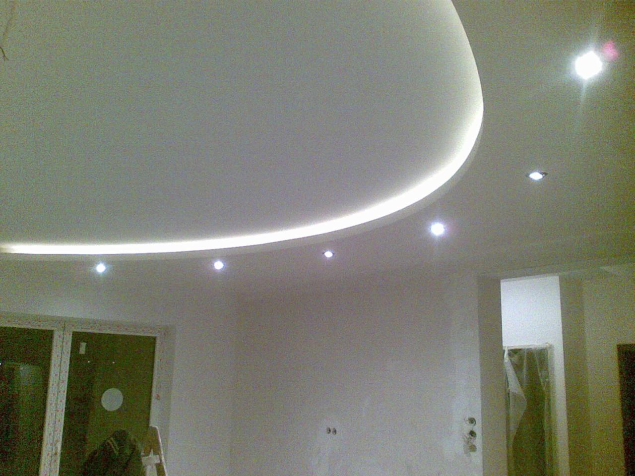 Nowoczesny sufit podwieszany z oświetleniem LED w kształcie łuku, punktowe lampy wkomponowane w gładką powierzchnię, widoczne okno i drzwi w tle.