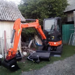 Pomarańczowa mini koparka Hitachi na trawiastym terenie, z dodatkowym osprzętem obok, w tle budynki gospodarcze i drzewa. Widok z boku, lekko z dołu.