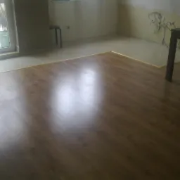 Układanie paneli podłogowych w pokoju z fragmentem przygotowanej do malowania ściany z wystającymi przewodami elektrycznymi, widoczny fragment stołu i okna.