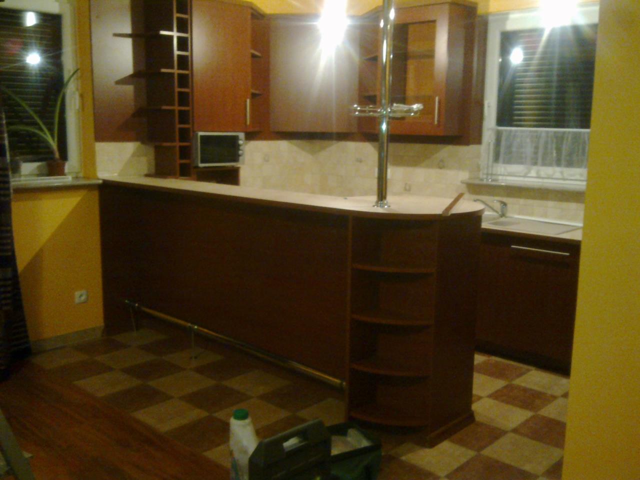 Wykonana na zamówienie kuchnia z barkiem, brązowe fronty szafek, metalowy reling pod blatem, szachownica na podłodze, widoczne narzędzia i butelka chemii gospodarczej.