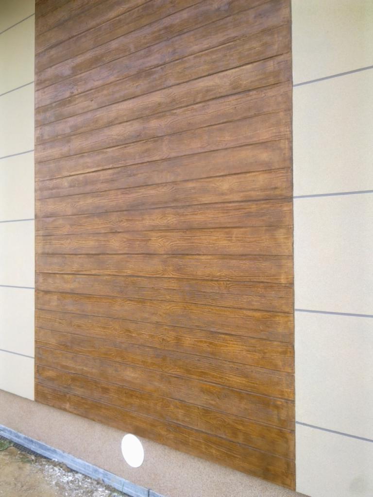 Fragment elewacji budynku z dekoracyjną, brązową okładziną imitującą drewniane deski, sąsiadującą z jasną, gładką powierzchnią z szarymi liniami podziału.