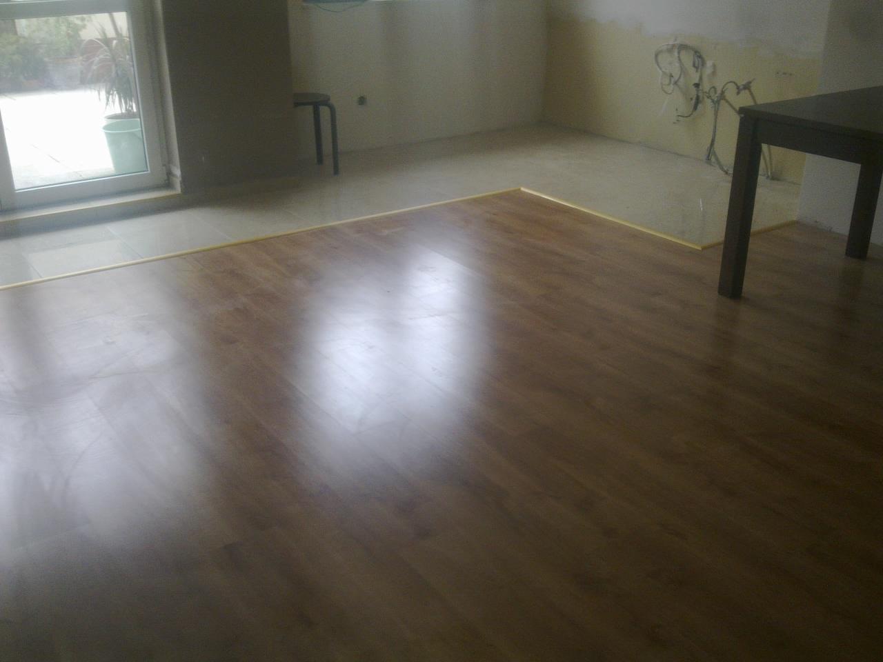 Układanie paneli podłogowych w pokoju z fragmentem przygotowanej do malowania ściany z wystającymi przewodami elektrycznymi, widoczny fragment stołu i okna.