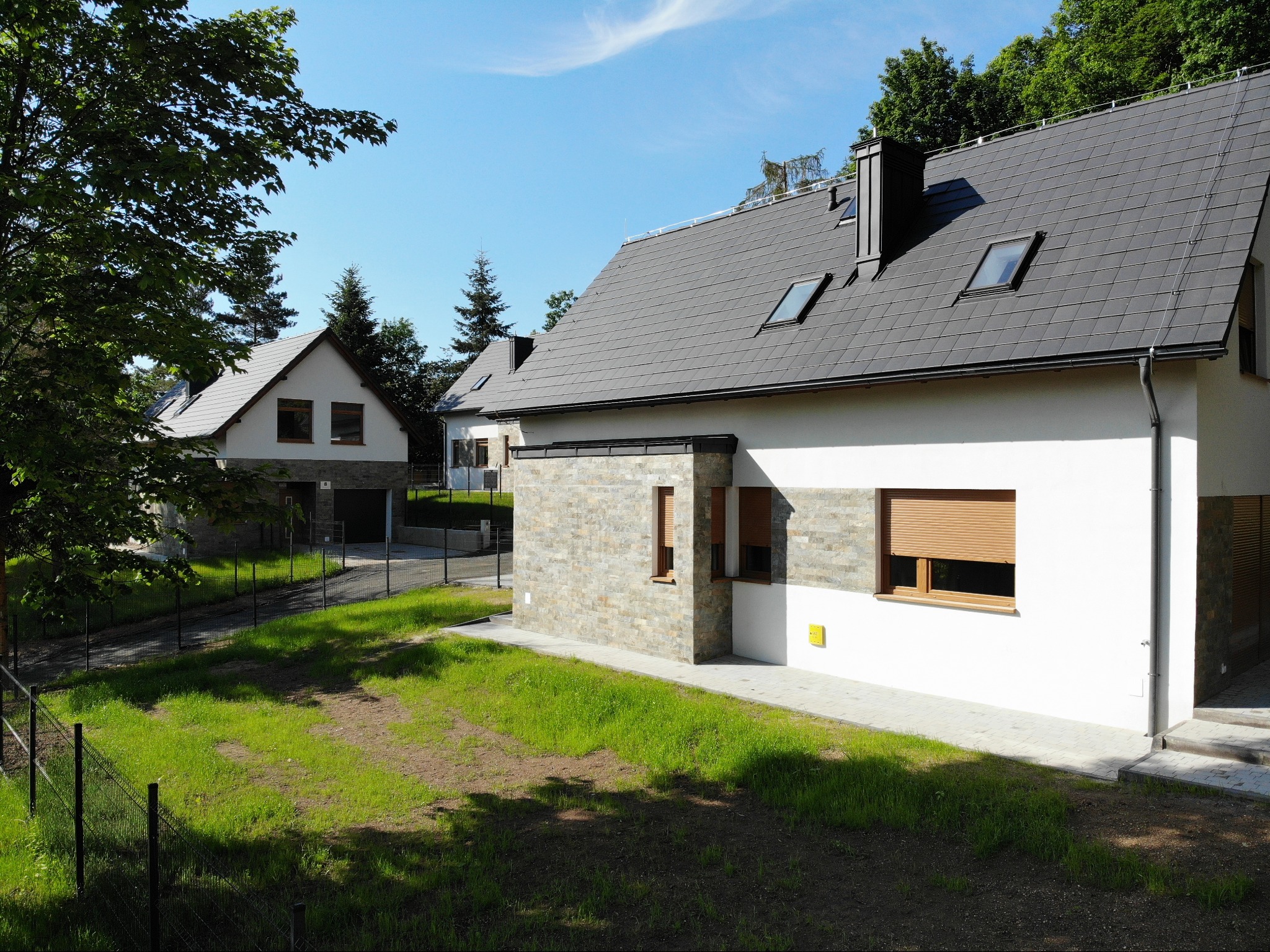 Nowoczesny dom z szarą dachówką i białymi ścianami, wykończony kamieniem, z oknami dachowymi i roletami zewnętrznymi, otoczony trawnikiem i ogrodzeniem.