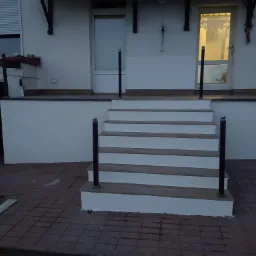 Wykończone schody zewnętrzne z płytkami imitującymi drewno i czarnymi balustradami, prowadzące do wejścia do domu z kostki brukowej.