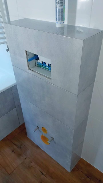 Obudowa stelaża podtynkowego WC wyłożona szarymi płytkami, widoczne przyłącza wody i otwór rewizyjny z niebieskimi i zielonymi elementami instalacji, tuba silikonu na górze obudowy.