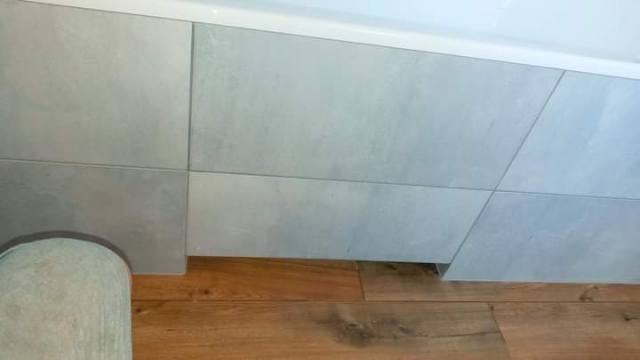 Obudowa wanny wykończona szarymi płytkami o matowej powierzchni, widoczna z perspektywy podłogi z paneli drewnopodobnych.