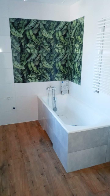 Nowoczesna łazienka z wanną obudowaną szarymi płytkami, białą glazurą i ścianą z motywem tropikalnych liści, drewnopodobna podłoga i białe żaluzje.