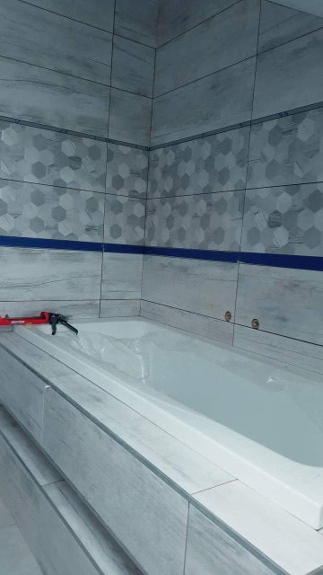 Wykończona łazienka w szaro-białych płytkach z motywem drewna i heksagonów, widoczna wanna zabezpieczona folią oraz aplikator do silikonu w rogu.