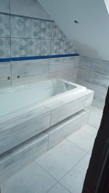 Wykończona łazienka na poddaszu ze skośnym sufitem, obłożona płytkami imitującymi drewno i mozaiką heksagonalną, wanna obudowana płytkami, zabezpieczona folią ochronną.