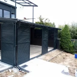 Zadaszony taras z przesuwnymi panelami, konstrukcja aluminiowa w kolorze antracytowym, częściowo otwarte panele, widoczny fragment domu z roletami zewnętrznymi w oknach oraz elementy ogrodu w tle.