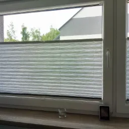 Szara, plisowana osłona okienna zamontowana w białym oknie PCV; na parapecie szklanka i elektroniczny termometr.