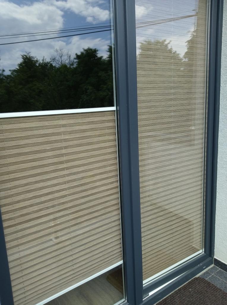 Beżowe plisy okienne zamontowane w oknie balkonowym z ciemną ramą, widoczne odbicie drzew w szybie.