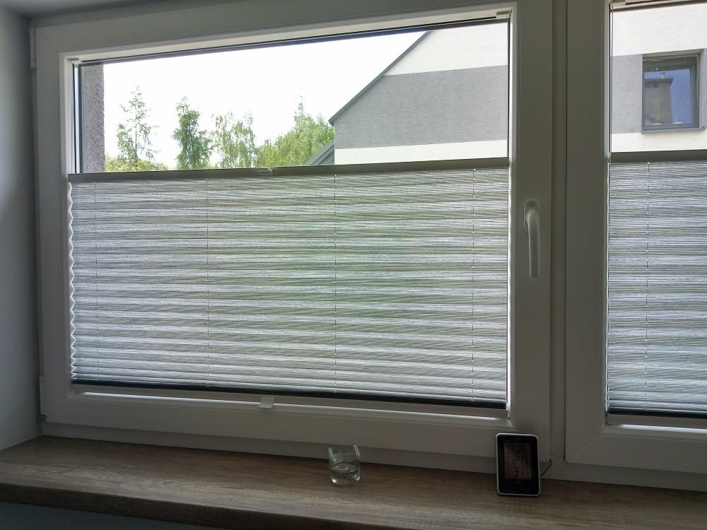 Szara, plisowana osłona okienna zamontowana w białym oknie PCV; na parapecie szklanka i elektroniczny termometr.