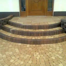 Półokrągłe schody z kostki brukowej w odcieniach beżu i brązu prowadzące do drewnianych drzwi wejściowych. Na podłożu wzór wachlarzowy.