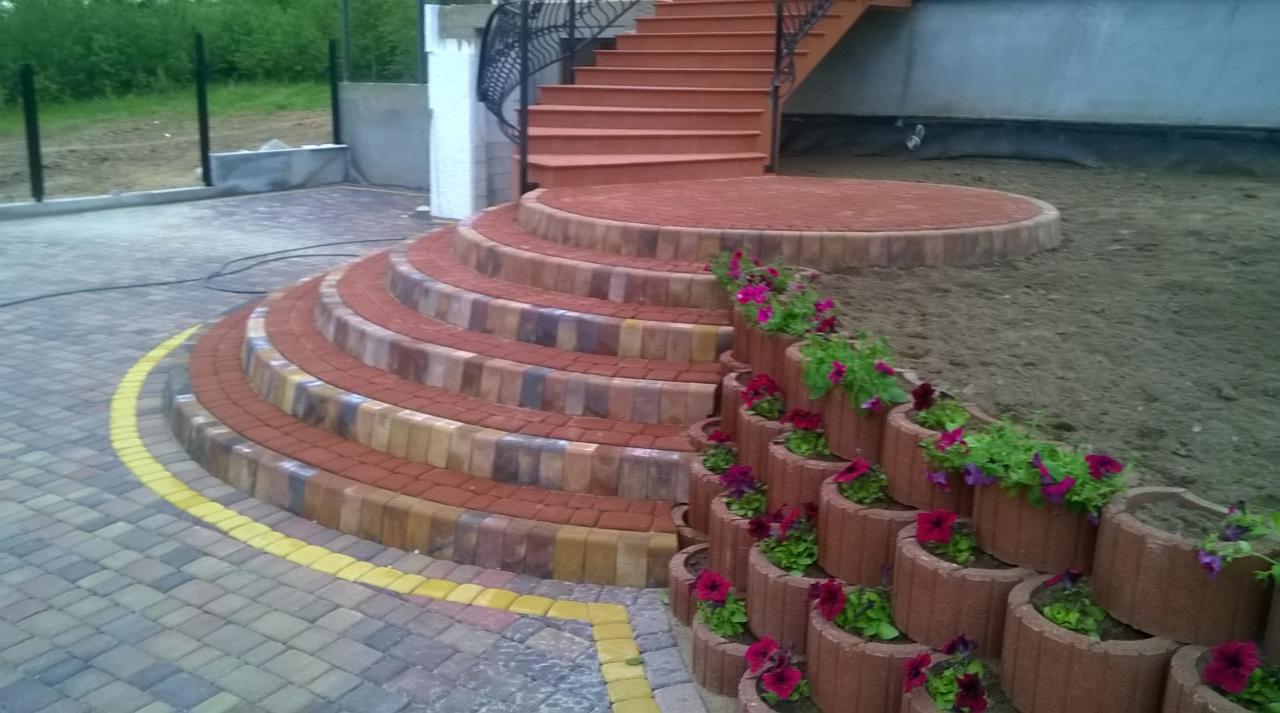 Kaskadowe schody z kostki brukowej prowadzące do góry, ozdobione donicami z kwiatami. Ułożona nawierzchnia z kostki brukowej w odcieniach szarości i żółci.