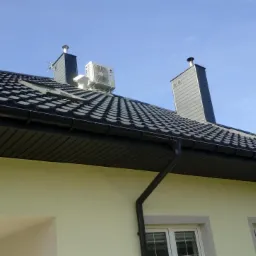 Zewnętrzna jednostka klimatyzacji zamontowana na dachu domu jednorodzinnego z czarną dach&oacute;wką, widoczna z poziomu gruntu, z dwoma kominami w tle.