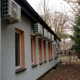 Rząd klimatyzatorów marki Gree zamontowanych na zewnętrznej ścianie budynku z oknami w pomarańczowych ramach, widok z boku na tle drzew bez liści i metalowego ogrodzenia.