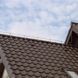 Brązowy dach z dachówki ceramicznej zabezpieczony instalacją odgromową z metalowymi masztami, na tle błękitnego nieba z chmurami.