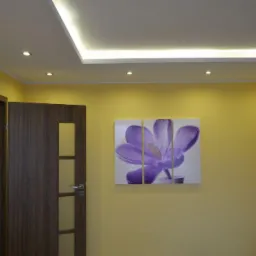 Żółta ściana w pokoju z obrazem fioletowego kwiatu, brązowe drzwi z przeszkleniami, sufit podwieszany z oświetleniem punktowym i lampą wiszącą.