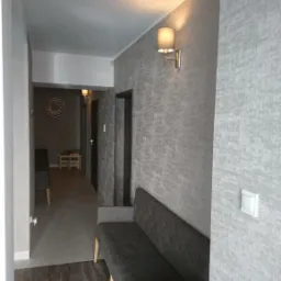 Szary korytarz z ławką obitą materiałem, lampą ścienną i podłogą imitującą drewno, widziany z perspektywy wejścia.