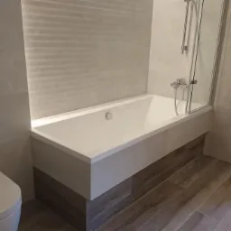 Nowoczesna łazienka z białą wanną obudowaną płytkami, szklanym parawanem nawannowym, grzejnikiem drabinkowym i podwieszaną toaletą z przyciskiem dwudzielnym, wykończona płytkami imitującymi drewno.