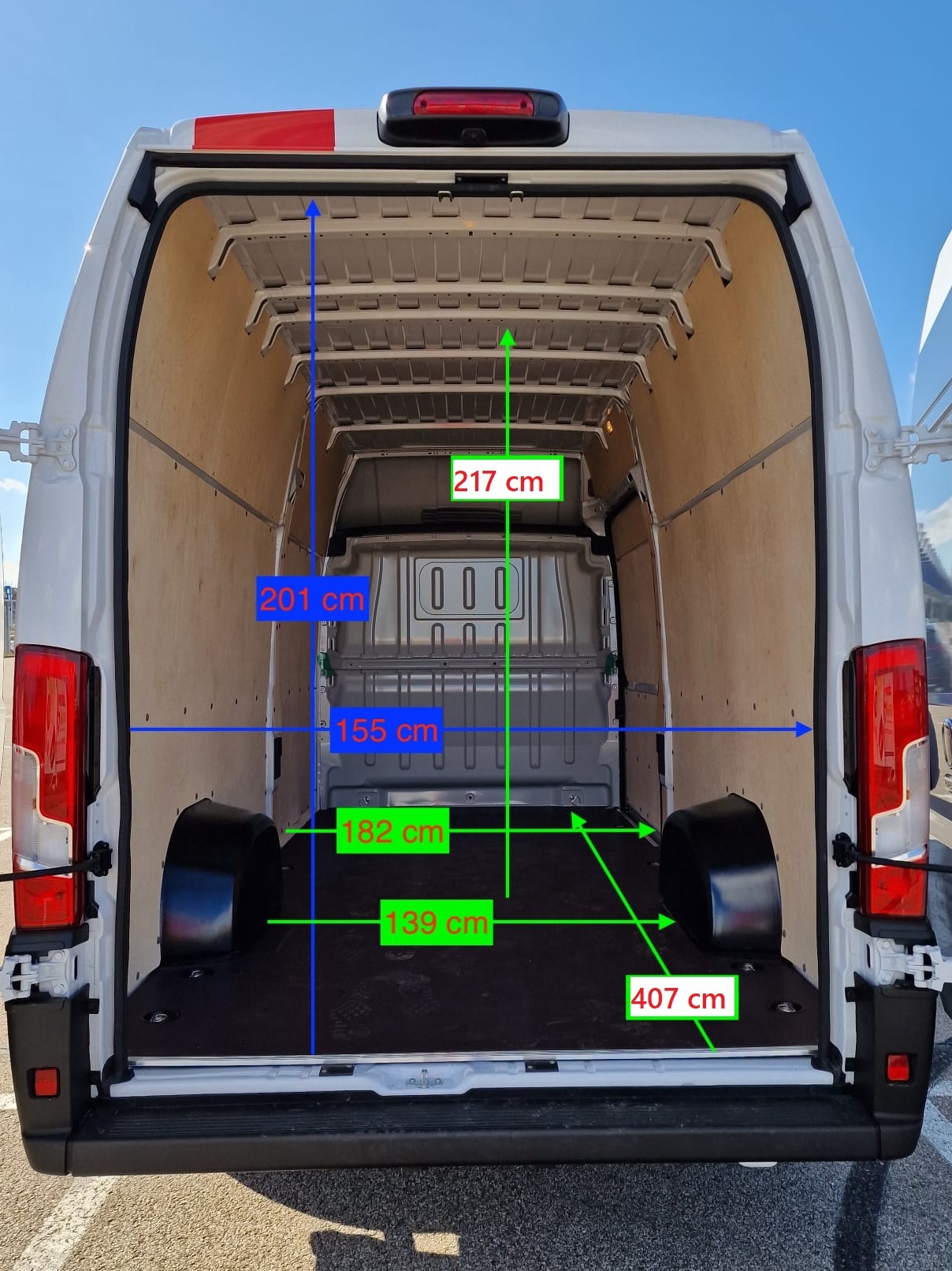 Wnętrze białego busa z otwartymi drzwiami, widok z tyłu, prezentacja wymiarów przestrzeni ładunkowej z zaznaczonymi odległościami w centymetrach.