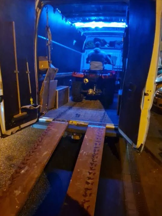 ATV na rampie wewnątrz oświetlonego na niebiesko furgonetki z otwartymi drzwiami, przygotowany do transportu. Widok z tyłu.